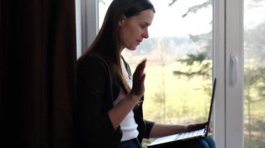 Mutlu genç bir kadın pencerenin pervazında oturmuş el sallıyor uzaktan video çekiyor online sohbet yapıyor dizüstü bilgisayarına bakıyor, gülümseyen kız internet bilgisayarıyla iletişim kuruyor.