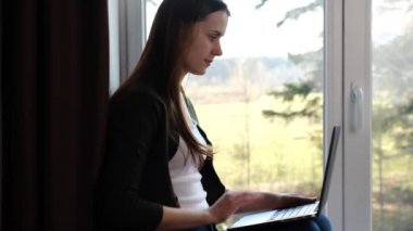 Mutlu genç bir kadın el sallıyor uzaktan video görüşmesi yapıyor çevrimiçi sohbet ediyor evdeki pencerenin pervazında dizüstü bilgisayarına bakıyor güzel bir kız gülüyor ve web kamerasıyla konuşuyor. Sosyal uzaklık