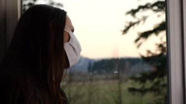 Steril yüz maskeli düşünceli bir kız pencere kenarına oturmuş üzgün üzgün bakıyor, depresif bir kadın evde yalnız vakit geçiriyor, karantina sırasında yalnız hisseden genç bir kadın Coronavirus salgını sırasında kendini yalnız hissediyor.