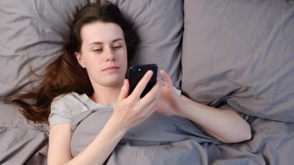 醒来时兴奋的年轻女子躺在床上 手里拿着智能手机欢快地尖叫着 从早晨开始的一天中得到了好消息 积极的情绪和幸福的概念 — 图库视频影像
