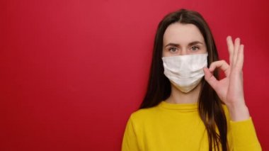Korunaklı beyaz tıbbi maskeli genç beyaz kadın, Coronavirus salgınına hazırlık yapıyor, kırmızı arka planda izole edilmiş, boş fotokopi alanında bir şeyler gösteriyor..