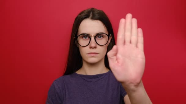 眼鏡の真面目な若い女性は 停止ジェスチャーでカメラに向かって手を引くと 落ち着いて 心配を停止するように求め 限界を示し 赤い背景に隔離された紫のTシャツを着ています — ストック動画
