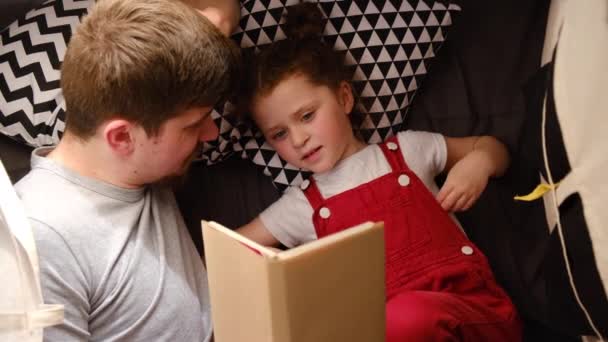 小さなかわいい娘を持つ幸せな若いお父さんのトップビュー読書本を保持します 笑顔のお父さんと子供の女の子は楽しい遊びを持っています 一緒に家で過ごす時間でテントに横たわっています 子供の読者の概念 — ストック動画