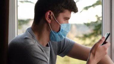 Tıbbi maske takan genç bir adam, elinde akıllı telefon, ekrana bakıyor, pandemik koronavirüsle ilgili kötü haberler alıyor. Erkek okuma nahoş e-posta, evde pencere pervazında oturan