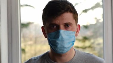 Evdeki pencere eşiğinde oturan, virüs maskesi takan genç bir adamın portresi salgın hastalık riskini düşünüyor ve kameraya üzgün bir şekilde bakıyor. Salgın, Coronavirüs 2019-ncov konseptini yayıyor.