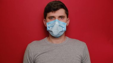 Steril yüz maskesi takmış ciddi bir genç adam dur hareketi yaparak kameraya doğru elini çekiyor. Sınırlarını gösteriyor. Kırmızı arka planda izole edilmiş gri tişört giyiyor. Karantina salgını koronavirüs virüsü konsepti