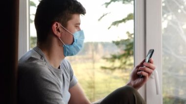 Oturma odasının pervazında oturan virüs maskesi takan bir erkeğin modern cep telefonu cihazıyla görüntülü konuşması akıllı telefon kullanarak internette selamlaşması. Coronavirus 2019-ncov konsepti
