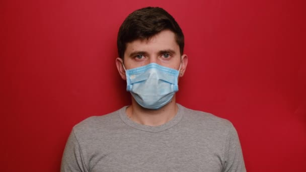 手を拒否すると 赤いスタジオの背景フェンスに隔離された保護医療マスクとグレーのTシャツの若い男性は カメラを見て不満男 流行病 コロナウイルス2019 Ncovコンセプト — ストック動画