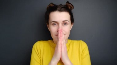 Çekici bir kadın dua etmek için umutla el ele tutuşuyor, yardım ve destek isteyen sevimli genç bir kız, kameraya bakıyor, sarı süveter giyiyor, gri stüdyo arka planında izole edilmiş.