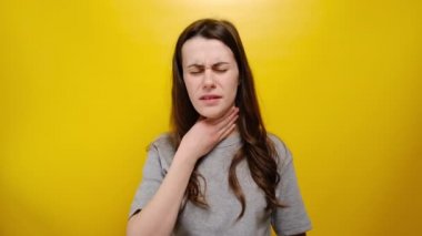 Mutsuz genç kadının boğazı ağrıyor, hasta ve iyi değil, boğazı ağrıyor, üşüttükten sonra acı çekiyor, gri tişört giyiyor, sarı arka planda duruyor. Rahatsızlık hastalıktan kaynaklanıyor.