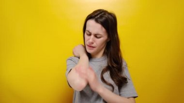 Mutsuz genç bir kadın dirsek ağrısı çekiyor ve masaj yapıyor. Hastalık sağlığı problemi, gri tişört giymiş, sarı arka planda izole edilmiş. Kol ağrısı ve yaralanma konsepti. Dirsek şikayeti olan kız.