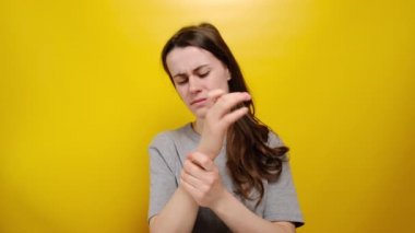 El veya bilek ağrısından muzdarip, gri tişört giymiş, sarı arka planda izole edilmiş genç bir kadın. Karpal tünel sendromu, artrit, nörolojik hastalık konsepti. Elin uyuşması.