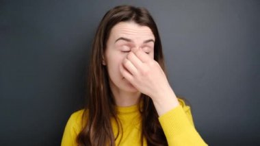 Acı gözler konsepti. Burun köprüsüne masaj yapan, göz yorgunluğu çeken, astenopi sorunu olan, mutsuz bakışlı, süveter giyen, gri arka planda izole edilmiş genç bir kadın..