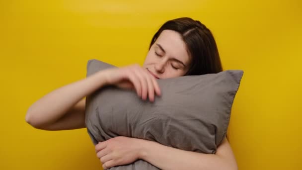 幸せな女性は灰色の枕で目を覚ます 頬に触れると 楽しくカメラを見て パジャマ姿で 健康的な睡眠の後に良い気分で 黄色の背景に隔離されて住んでいます 眠りの概念 — ストック動画