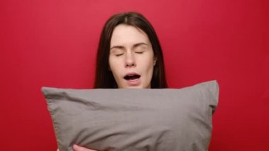 Uykulu mutlu genç kadın esniyor gri yastığı kucaklıyor, uyumak istiyor, pijama giyiyor, kırmızı arka planda poz veriyor ve terfi için boş bir alan yaratıyor. Uyku konsepti