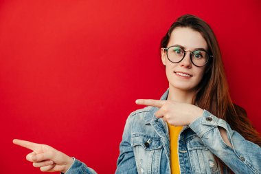 Gözlüklü güzel esmer kadın, elleri bir yana, reklam ya da promosyon metnin için fotokopi alanı gösteriyor, kot ceket giymiş, kırmızı arka planda izole edilmiş. Tanıtım konsepti