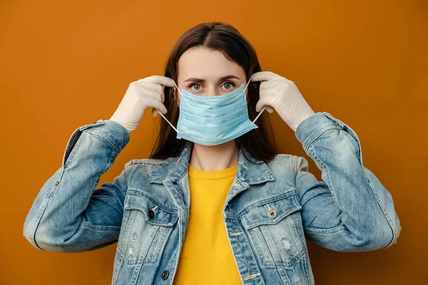 Woman wears protective medical mask not to infect other people, has virus infection, wears denim jacket, poses indoor against brown wall. Epidemic pandemic spreading coronavirus 2019-ncov concept