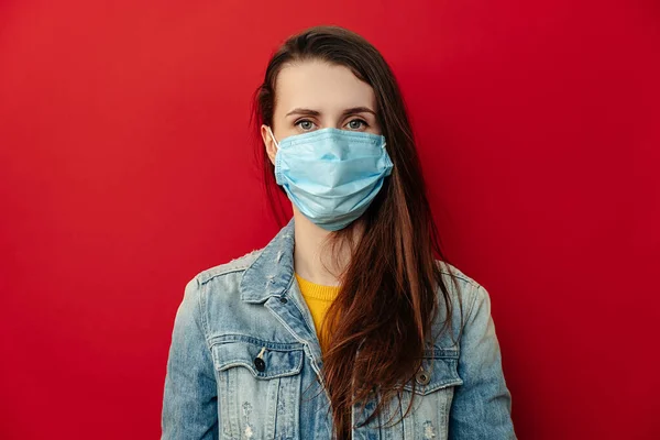 Serious young woman wears respirator mask, looking confident at camera, wears denim jacket, isolated against red background. Epidemic pandemic spreading coronavirus 2019-ncov concept