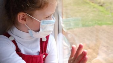 Üzgün küçük bir kızın pencereye bakıp sorunları düşünmesi, tıbbi maske takan üzgün bir çocuğun salgın hastalık riskini düşünmesi. Evde kalıp koronavirüs salgınını önleyin.