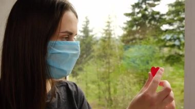 Hastalıkla mücadelede doktorlara ve hemşirelere şükranlarını göstermek için yüzü kırmızı pencereye yakın tıbbi maske takan hoş bir kadın portresi. Covid-19. Seçici odak