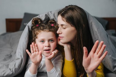 Şirin küçük çocuk kızı ve neşe dolu genç anne battaniyeyle kaplı bir şekilde yatağa uzanıyor. Uzaktan telefon açıp video bloğu çekiyor. Kameraya bakıyor, web kamerasına bakıyor, gülüyor, evde eğleniyor.