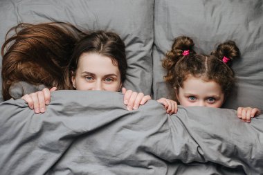 Sevimli komik çocuk ve genç anne portresi yumuşak bir battaniyenin altına saklanır, yatakta eğlenir, kameraya bakar, günaydın der. Gülümseyen anne ve küçük kız eğlenin.