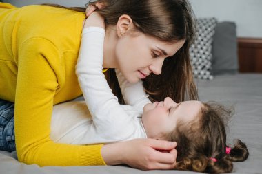 Anaokulundaki küçük kızın rahat yatağında yatıp gülümseyen bir anneyle oynaması, neşeli bir annenin tatlı küçük kızını kucaklaması, gülmesi, yatak odasında hafta sonunun tadını çıkarması, birlikte eğlenmesi.