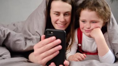 Mutlu genç bir kadın battaniyeyle örtülmüş bir şekilde yatarken tatlı, gülümseyen küçük bir kız çocuğuyla, akıllı telefondan selfie çekerken. Yatak odasında mobil uygulamalar kullanan neşeli bir aile.