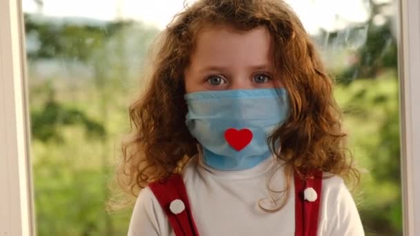 可爱的小女孩戴着红心的面部医疗面具 快乐有趣的学龄前幼儿坐在窗台上看着相机 Coronavirus或Covid 19流行病症状 — 图库视频影像
