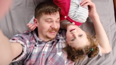 Küçük kızının yatakta uzanmış, kamerada konuşurken gülümseyen web kamerasıyla çevrimiçi video konuşması yaptığı neşeli bir baba manzarası. Baba ve küçük kız rahat yatak odasında rahatlayın.