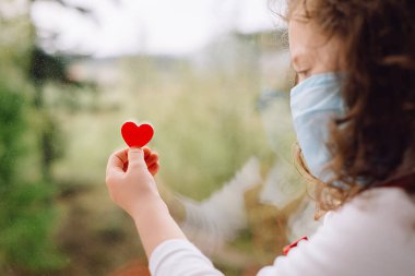 Koruyucu yüz maskesi takan, elinde küçük kırmızı bir kalple evde oturan okul öncesi küçük kızın yan görüntüsü. Evde kalıp koronavirüs salgınını önleyin. Ev karantinası