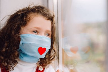 Koruyucu yüz maskesi takan, kırmızı kalpli, pencerenin pervazında oturan, bulanık pencereye bakan okul öncesi kızı. Kavram, Covid-19 salgınındaki tüm önemli çalışanlara teşekkür et