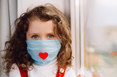 Evde pencere eşiğinde oturan, kırmızı kalpli virüs maskesi takan anaokulu çocuğu, küçük kız kameraya bakıyor. Salgın, Coronavirus 2019-ncov 'u yayıyor. Hemşire günü konsepti.