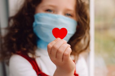 Koruyucu tıp maskeleri takmış sevimli bir anaokulu kızı elinde kırmızı bir kalple Covid-19 salgınındaki tüm önemli çalışanlara teşekkür ediyor. Seçici odak