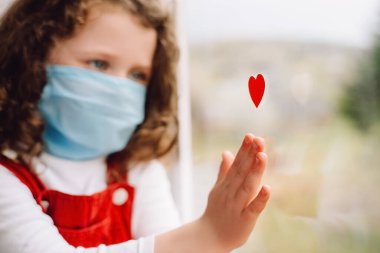 Hastalıkla mücadelede doktorlara ve hemşirelere şükranlarını göstermek için kırmızı kalpli, tıbbi maske takan küçük şirin bir kız portresi. Covid-19. Seçici odak