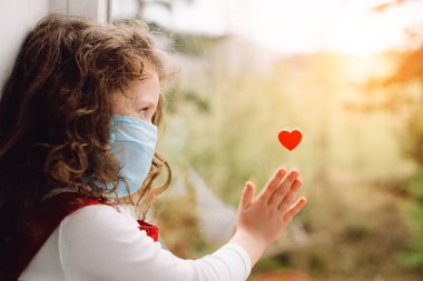 Mavi tıbbi maske takan küçük bir kız hastalıkla mücadelede doktorlara ve hemşirelere teşekkür etmek için penceresinde küçük kırmızı bir kalple oturuyordu. Covid 19