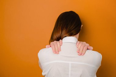Kahverengi stüdyo arka plan masajında izole edilmiş beyaz tişörtlü genç esmer kadına yakından bakın. Kas spazmından muzdarip, sırtı ağrıyan sağlıksız bir kızın boynuna dokunması veya aniden incinmesi.