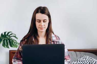 Genç, güzel, serbest çalışan bir bayanın dizüstü bilgisayardan e-posta yazarken, bilgisayarla evde rahat bir yatakta otururken, internetten ders çalışırken ön görüntüsü..