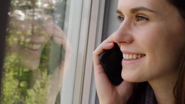 快乐美丽的年轻女性在家里用智能手机交谈 微笑可爱的女孩用手机接听电话 美丽的女士用手机坐在窗台上愉快地交谈 — 图库视频影像