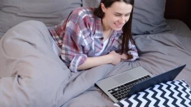 Mutlu genç bir kadın, battaniyenin altında rahat bir yatakta uzanıp uzaktan video görüşmesi yapıyor. İnternetten sohbet ediyor, dizüstü bilgisayarına bakıyor, güzel bir kız web kamerasına gülüyor ve iletişim kuruyor. Sosyal uzaklık