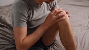 Evde oturmuş, dizine elleriyle dokunan, acı hisseden mutsuz genç bir adamın yan görüntüsü. Bacağı ağrıyan sakallı adam, kendini iyi hissetmiyor, sağlık problemi kavramı, osteoartrit