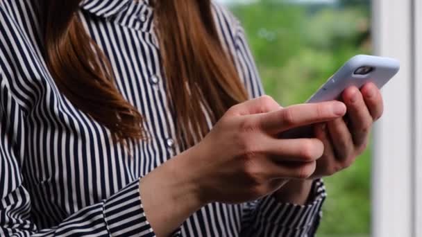 年轻的千禧年女孩或妇女站在窗边 手持智能手机应用的概念 在线搜索信息 查看社交网络 在手机上下订单的侧面照片 — 图库视频影像