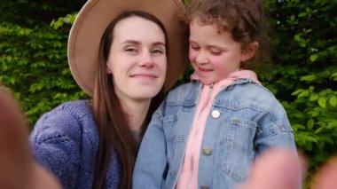 Şapkalı mutlu bir anne ve el sallayan sevimli bir kız çocuğu. Kameraya bakıyor. Yaz parkında video görüşmesi yapıyor.