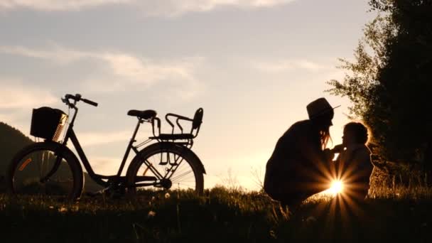 帽子の若い母親を愛し かわいい就学前の娘が自転車の近くに座って鼻に触れ 屋外で日没を楽しんでいます 家族関係と夏の野外活動の概念 — ストック動画