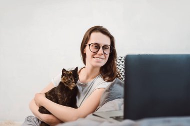 Siyah kedileriyle gözlüklü neşeli genç bir kadın dizüstü bilgisayar ekranına bakıyor, komik filmler izliyor ya da arkadaşlarıyla internette sohbet ediyor. Çekici kız evde bilgisayar kullanıyor.