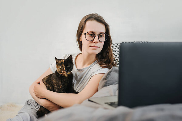 Cute caucasian young woman in eyeglasses relaxing resting on comfortable bed with cute pet cat watching remote education webinar class, movie series on laptop in cozy living room. Social distance