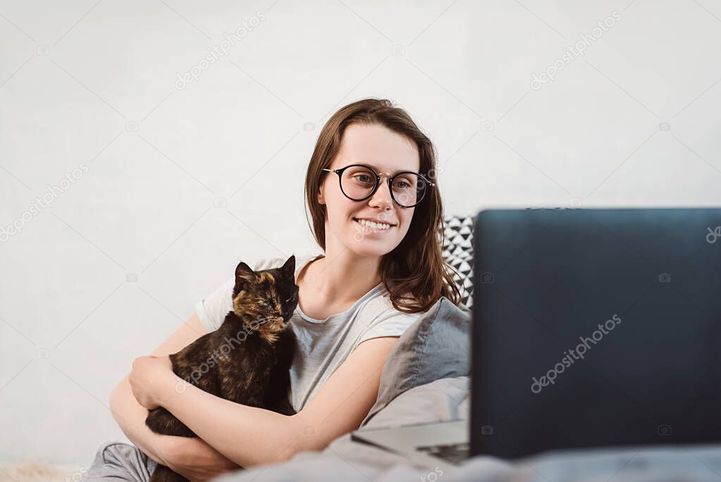 Cheerful smiling young woman in eyeglasses with her black cats looking at laptop screen, watching funny movies or chatting on internet with friends. Attractive girl uses a pc at home