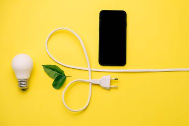 Enerji tasarrufu kavramı. LED ampulün, elektrik prizinin, küçük yeşil yaprakların ve sarı arkaplanda kopyalama alanı olan modern akıllı telefon modelleme bileşiminin yaratıcı üst görünümü düz yatağı