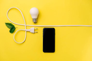 Çevre, biyoloji, enerji tasarrufu kavramı. LED ampul, elektrik prizi, küçük yapraklar ve en az sarı arka planda kopya alanı olan modern akıllı telefon kompozisyonu. Düz yatış, üst görünüm