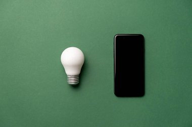 Yeşil zemin üzerinde fotokopi alanı olan LED ampulün ve modern telefon modelleme bileşiminin yaratıcı üst görünüm düz yatağı. Enerji tasarrufu, çevre dostu, küresel ısınma kavramı. Asgari biçim.